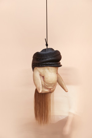 A surreal sculpture hanging from a cord features a human torso with a tire wrapped around it. The torso has long, reddish hair extending from its bottom, set against a light peach background.