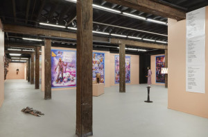 An art gallery with wooden beams and peach-colored walls features colorful posters and sculptures. The space is lit with overhead lights, and there are art pieces on both walls and pedestals.