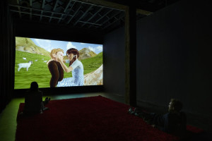 Two people sit on a red carpet in a dark room watching a large screen displaying an animated scene of two characters embracing in a green meadow with goats and mountains in the background.