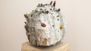 A textured ceramic sculpture with a rounded shape, covered in a mix of white, gray, and brown glazes, adorned with protruding sharp crystals of varying colors, displayed on a wooden pedestal.