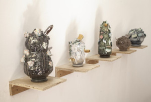 A row of five ceramic sculptures of varying shapes and textures, each on its own small wooden shelf mounted on a wall. The sculptures are decorated with a mix of colorful materials and glazes.