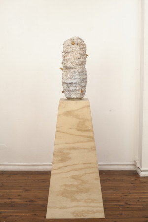 A tall, cylindrical ceramic sculpture with a rough, textured surface in shades of white and beige, with small golden elements, placed on a tall wooden pedestal in a gallery setting.