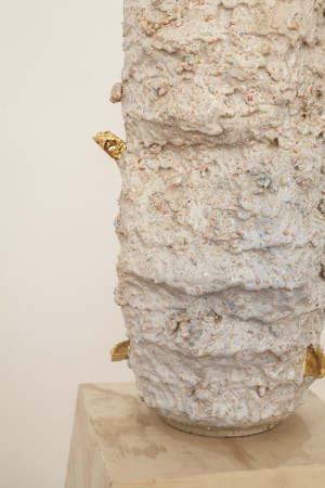 A close-up of the tall, cylindrical ceramic sculpture, showcasing its rough, textured surface with small, colorful specks and several small golden elements embedded in the sculpture.