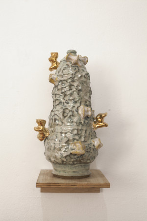 A textured ceramic sculpture with a tall, conical shape, decorated with a rough surface in shades of gray and beige, featuring several golden protrusions, displayed on a small wooden shelf.