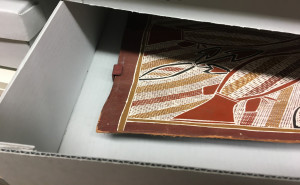 A photograph showing a corner of a bark painting artwork peeking out from a storage shelf.