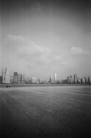 A black-and-white photograph shows a vast, empty beach with the skyline of Dubai in the background. The beach is wide and stretches into the distance, with a few scattered people visible, emphasising the openness of the space. The skyline features a mix of high-rise buildings and skyscrapers, creating a stark contrast between the natural sandy foreground and the urban landscape. The sky is partly cloudy, and the overall scene captures a calm and expansive view of the beach against the backdrop of the city.