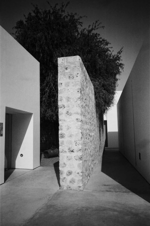 A black-and-white photograph shows a narrow corridor formed by a tall, rough-textured stone wall and smooth, modern white walls on either side. The stone wall extends vertically, creating a stark contrast with the sleek surfaces of the adjacent structures. In the background, dense foliage is partially visible, casting shadows on the ground. The scene captures the interplay between traditional and contemporary architectural elements, emphasizing texture and form.