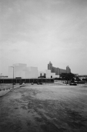 A black-and-white photograph shows the exterior of the Jameel Art Centre in Dubai, UAE. The foreground features a large, empty, and slightly dusty open area leading to a parking lot with a few parked cars. The art centre itself is characterised by its modern, geometric architecture with white cubic structures. In the background, tall, sleek buildings rise, adding a touch of urban landscape to the scene. Palm trees are visible to the right, enhancing the contrast between the natural and built environments. The sky is overcast, contributing to the photographer's contemplative mood.