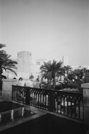 A black-and-white photograph captures a scene at Madinat Jumeirah in Dubai, UAE. The foreground features a decorative railing with gothic-style arches, overlooking a small fountain with water jets. Beyond the railing, palm trees and lush landscaping frame the view of traditional Arabian-style buildings with wind towers and crenellated parapets. People can be seen strolling through the area, enjoying the outdoor space. The architecture and landscaping evoke a blend of historic and modern aesthetics, creating a picturesque and tranquil atmosphere.