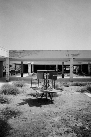 A black-and-white photograph shows an abandoned kindergarten in Kalba, Sharjah, UAE. In the foreground, an old, rusted merry-go-round with seats is positioned in a barren, dirt-covered playground with sparse, overgrown vegetation. The background reveals a dilapidated, open-air structure with peeling paint and graffiti on the walls. The scene evokes a sense of neglect and decay, capturing the remnants of a once lively and active place for children.