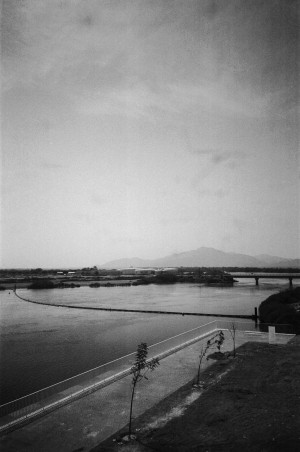 A black-and-white photograph depicts a serene landscape featuring a body of water with a gentle curve, bordered by a paved walkway with a railing on the left side. Sparse young trees line the walkway. In the background, there is a bridge crossing the water, leading to a flat expanse of land with a few buildings and vegetation. Distant mountains are faintly visible through the hazy sky, adding depth to the tranquil scene. The image captures a quiet moment with a focus on the natural and constructed elements of the environment.