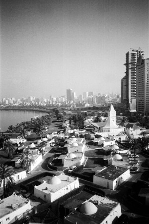 A black-and-white photograph depicts a cityscape with a mix of traditional and modern architecture. In the foreground, there are several low-rise buildings with domed roofs, interspersed with palm trees and greenery. To the right, a taller, unfinished high-rise building stands prominently. The background features a skyline of high-rise buildings along a waterfront, showcasing the blend of contemporary and traditional elements in the city. The image captures the architectural diversity and urban development of the area.