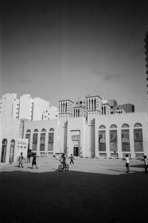 A black-and-white photograph captures a street scene in front of the Sharjah Art Museum. The museum building is prominent with its tall arched windows and large banners displaying exhibition information. In the foreground, several children are playing, with one riding a bicycle and others walking or standing around. The surrounding area includes additional buildings with traditional architectural elements, such as wind towers, blending with modern high-rise structures. The image captures a moment of everyday life against the backdrop of cultural and architectural heritage.