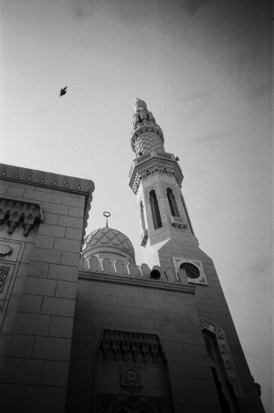 A black-and-white photograph captures a dramatic upward view of the Jumeirah Mosque in Dubai, UAE. The image focuses on the mosque's ornate minaret, which features intricate geometric patterns and arched windows. Adjacent to the minaret, a dome with a crescent moon finial is visible, along with part of the mosque's facade adorned with decorative details. A single bird is seen flying in the sky above, adding a sense of movement and life to the scene. The photograph emphasises the architectural beauty and spiritual significance of the mosque against a clear sky.