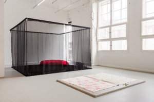 An art installation with a sheer black canopy over a red pillow and adjacent large paper with pink and blue markings on the floor of a bright room with large windows.