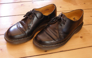 A pair of well-worn, black leather lace-up shoes with yellow stitching on a wooden floor.