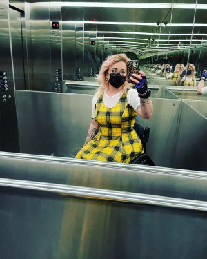 Robin, a white wheelchair user with pink hair and a mask, taking a selfie in a lift.