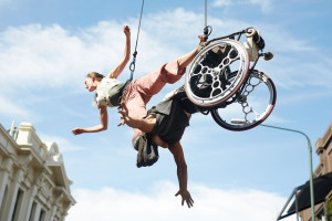 A previous iteration of The Air Between Us (2021). Rodney Bell and Chloe Loftus are suspended from rigging. Bell is tilted backwards, arms outspread, while Loftus has one foot hooked around his neck and the other resting on the frame of his wheelchair.